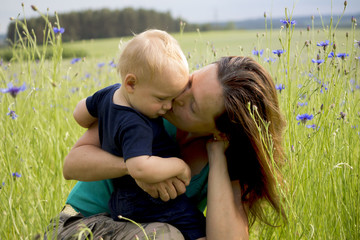 Happy family in the field of blew flowers. Beautiful young mother kisses her adorable baby boy. Concept of happiness
