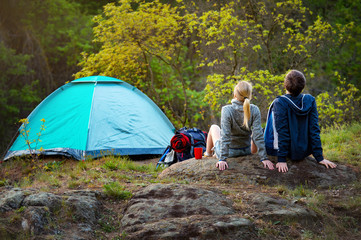 Young couple hiking with backpacks resting near the tent in the warm sunshine at sunset. Travel, vacation, holidays and adventure concept. Forest Mountain landscape background