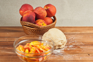 Organic ingredients and preparations for homemade peach pie with a basket of whole peaches and a clear glass bowl of sliced peaches and a ball of raised dough on wood cutting board