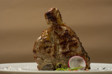 Succulent grilled large t-bone steak garnished with herbs, radish and salt, on a white plate; brown...