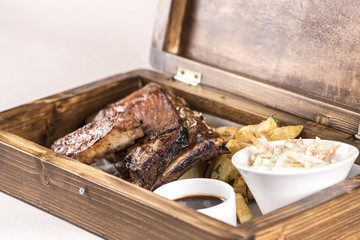 Fancy dish with grilled pork ribs, with barbecue sauce, French fries decorated with radish and red and green leafs, Coleslaw salad, all served in a wooden box, light background, isolated