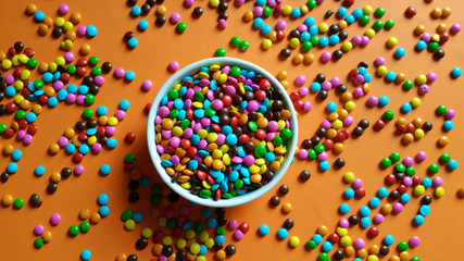 Colorful candy confetti in white pot and scattered over orange background top view
