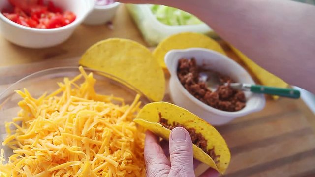 A man spoons ground beef into a taco shell with grated cheese, chopped tomatoes, onions and lettuce on the cutting board beneath