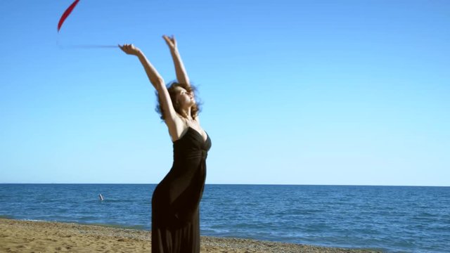 A young woman in a black dress is standing on the sand and doing a drawing of a red ginastic ribbon on the background of the sea and sky.