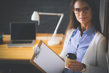 Young woman standing near desk with laptop holding folder and cup of coffee. Workplace. Business...