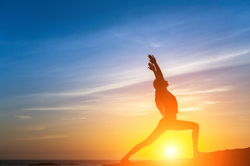 Silhouette of young fitness woman doing exercises on the sea beach during sunset. Yoga and healthy...