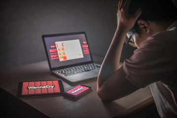 Young Asian male frustrated, confused and headache by WannaCry ransomware attack on desktop screen, notebook and smartphone, cyber attack internet security concept