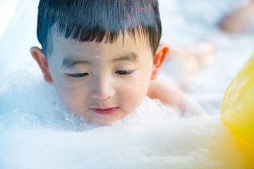 Asian boy playing with water and foam in inflatable baby pool on hot summer day.