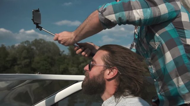 Slowmotion bearded man in sunglasses driving boat with friends on background peoples taking selfie with smartphone mounted on selfie stick