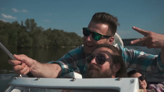 Slowmotion of sideview bearded man in sunglasses driving boat with friends on background peoples taking selfie with smartphone mounted on selfie stick