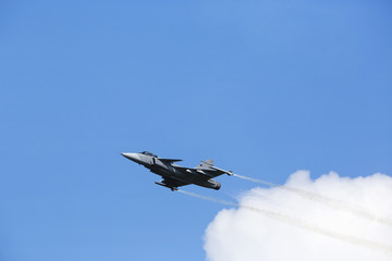 Modern tactical jet fighter is flying slowly with white smoke trail. Blue sky with clouds in the background.