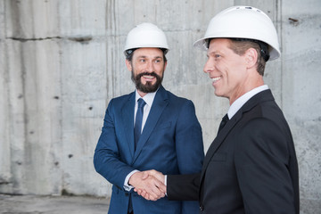 Professional mature architects in helmets shaking hands and smiling