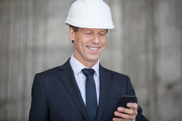 Smiling middle aged businessman in hard hat using smartphone
