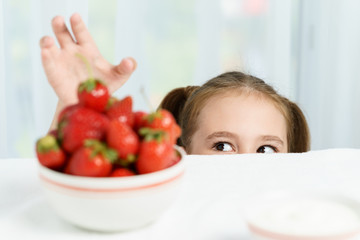 Young cute smiling european little girl is trying to steal ripe jucy strawberry from plate of many berries while she sitting under white table