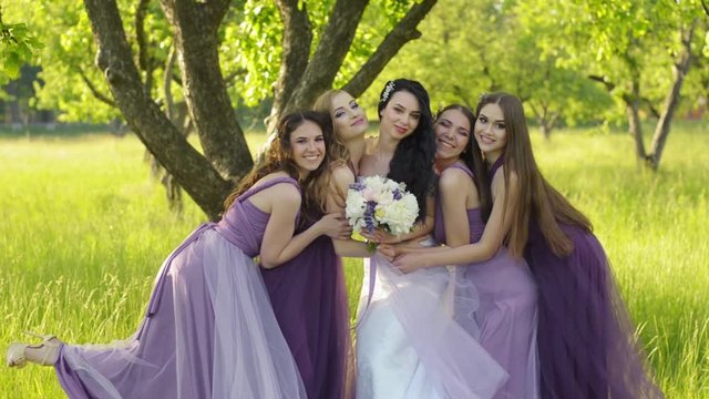 Emotional bridesmaids hugging the bride. Caucasian girls in purple wedding dresses smiling and posing outdoors.