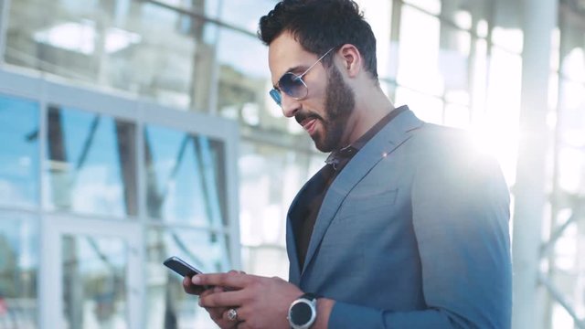 Handsome bearded Eastern businessman standing by the airport entrance, using his phone and smiles to received message, texts back. Stylish suit, sunglasses, elite watch. Successful lifestyle.
