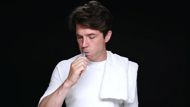 Unhappy brunette man with towel on shoulder brushing teeth isolated over black