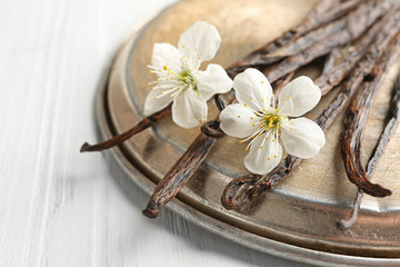 Dried vanilla sticks, flowers and metal plate on light wooden background, closeup