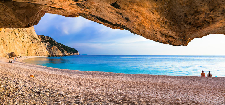 High resolution panorama of a romantic couple sitting on Porto Katsiki beach at sunset, view from inside a cave. Natural stone frame, white cliffs and turquoise sea in Lefkada, Greece.
