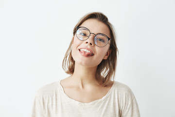Obraz premium Cute young pretty girl in glasses smiling showing tongue looking at camera over white background.