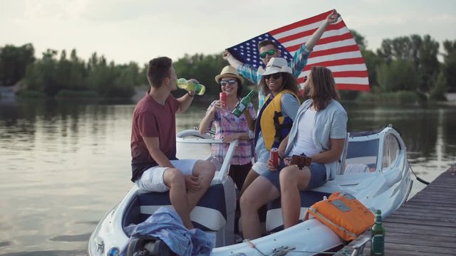 Group of cheerful friends having drinks on pier near boat celebrating Independence day and make selfie with smartphone mounted on monopod