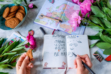 Summer planning holiday background - female hands with notebook, map, envelopes and postcards, peonies and cookies. Vacation planning concept. Top view, selective focus