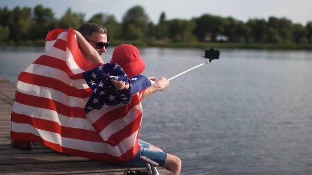 Slow motion of son raise American flag while his dad make self picture using cell phone mounted on monopod or selfie stick