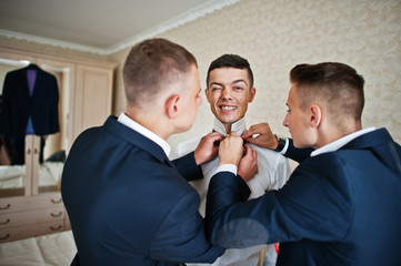 Groomsmen helping groom to dress up and get ready for his wedding in a room.