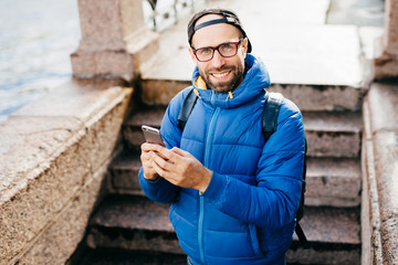 Young bearded guy in black cap and blue anorak holding rucksack standing outdoors with cell phone isolated over steps background. Stylish blogger with smartphone. People and lifestyle concept