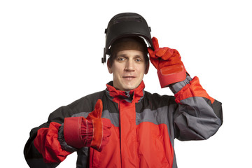 The man in working clothes and a welder mask shows a thumb and smiles..