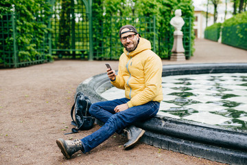 Obraz premium Smiling happy man with bristle wearing casual clothes and big eyewear sitting crossed legs near fountain in park holding cell phone messaging with his girlfriend having delightful expression