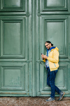 Stylish Man In Black Cap And Yellow Anorak Standing Sideways Against Green Background Holding Cell Phone Reading Funny Posts On His Social Network Page. People, Leisure, Technology, Style Concept