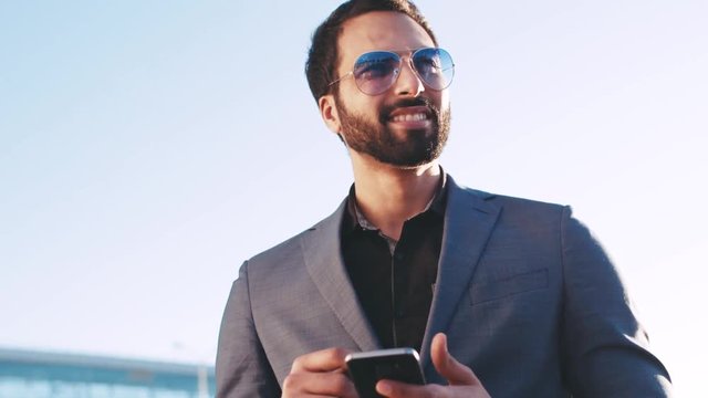 Smiling handsome young businessman in a formal suit using his phone, sliding the touchscreen while waiting for someone by the airport or office entrance. Online apps, internet surfing.