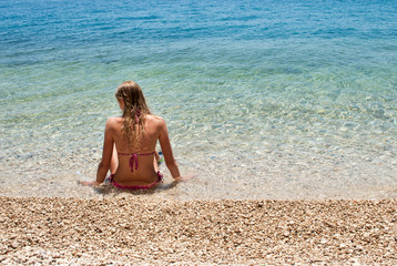 Fototapeta na wymiar Young blond female sitting at the edge of a calm shallow blue sea, looking to the left 