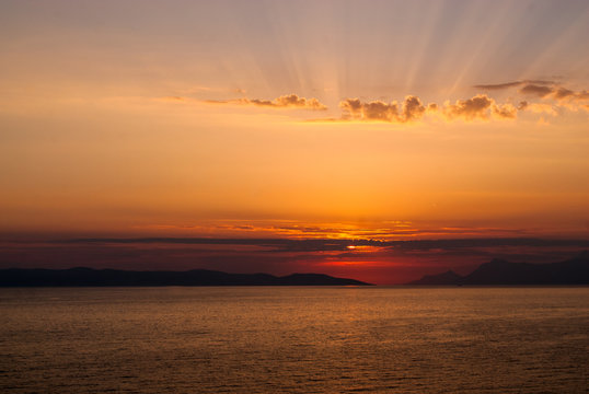 Horizontal image of golden sunset with sun low above the sea covered by clouds and sunrays coming through clouds above
