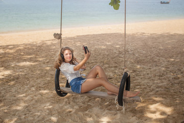 Girl sitting on the swing on the tropical beach, paradise island. Makes selfie