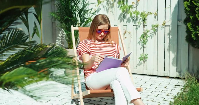 Young woman in sunglasses sitting on the beach chair in the garden and using reading book