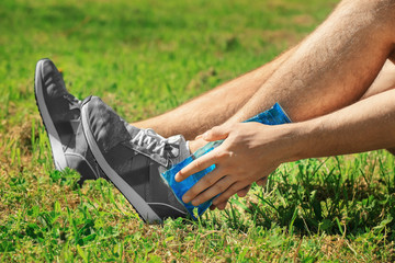 Young man applying cold compress to leg while sitting on grass outdoors, closeup
