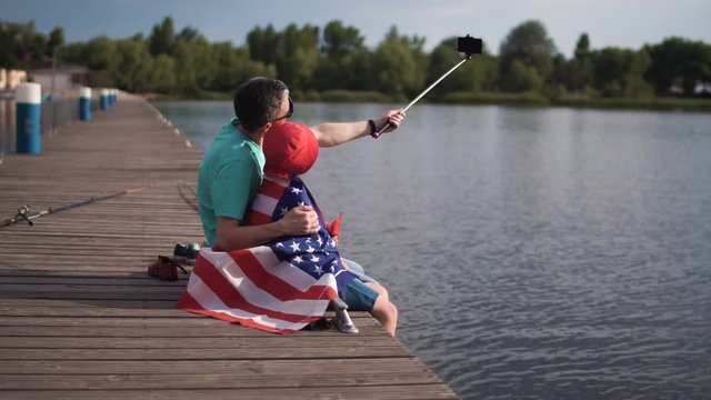 Mature man and boy sitting on pier and holding American flag while laughing and grimacing and make self photo using smartphone mounted on selfie stick