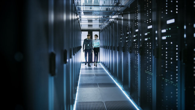 Female IT Technician and Male Server Engineer Talk and Discuss. They are in Working Data Center full of Rack Servers. Woman Holds Laptop.