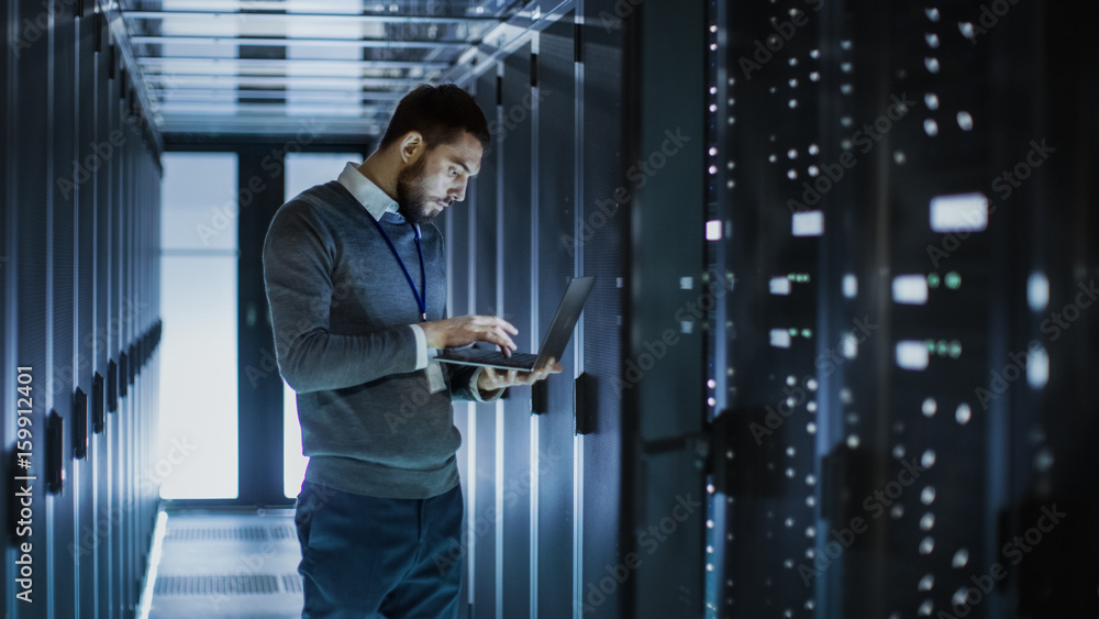 Poster it technician works on laptop next to a server cabinet in big data center. he runs diagnostics and m