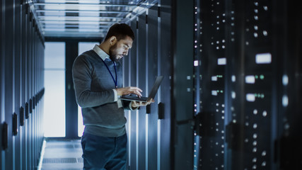 IT Technician Works on Laptop next to a Server Cabinet in Big Data Center. He Runs Diagnostics and...