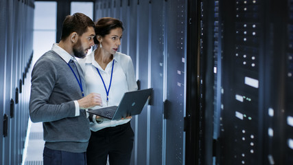 Male IT Specialist Holds Laptop and Discusses Work with Female Server Technician. They're Standing in Data Center, Rack Server Cabinet is Open. - obrazy, fototapety, plakaty