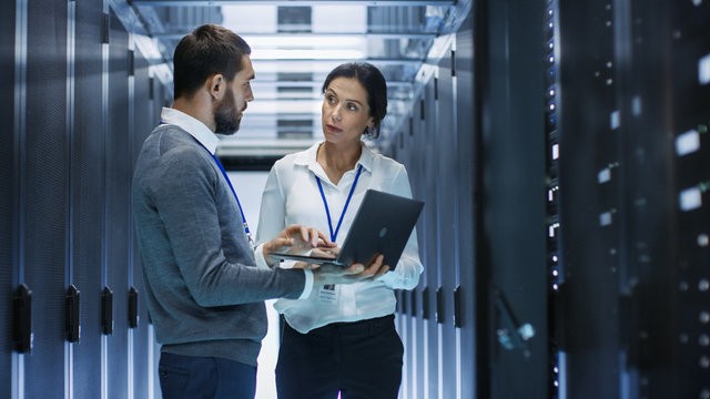 Male IT Specialist Holds Laptop and Discusses Work with Female Server Technician. They're Standing in Data Center, Rack Server Cabinet is Open.