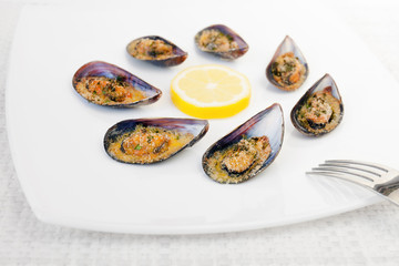 Gratin mussels. Grilled mussels cooked in the oven, accompanied by slices of lemon, grated pan and parsley. Mussels cooked on white plate.