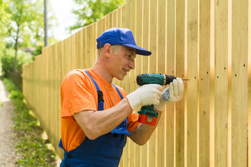 Handyman twirls a screw into the wooden fence wearily smiling