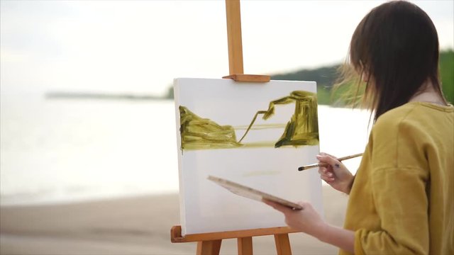 Young woman painter drawing watercolor landscape picture on easel. Beach of the lake