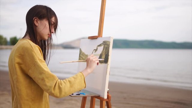 Young woman painter performing picture of landscape on the bank of the river. Countryside on background