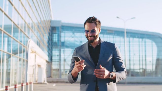 Attractive businessman walks by the airport, gets the message, uses the phone, is surprised with the result, reacts happily to it. Being a winner, happy emotions. True emotions.