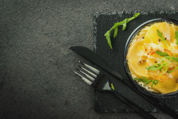 Casserole. French cuisine. Homemade potato gratin in a bowl for baking. On a black stone background. With leaves of fresh arugula for decoration. Top view copy space